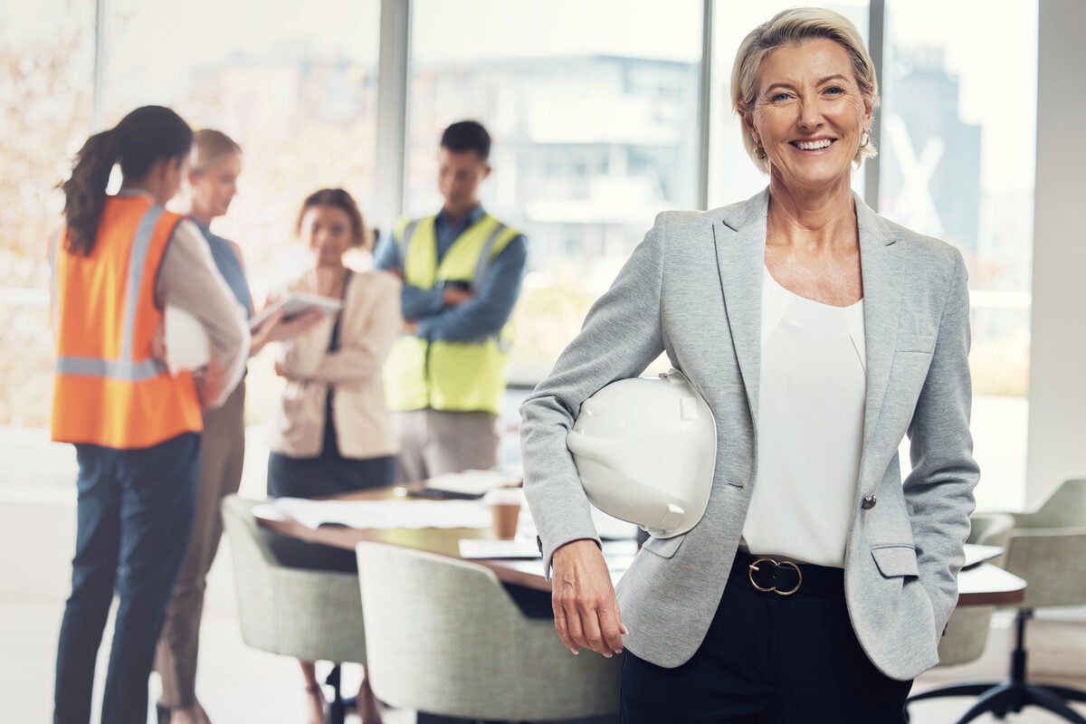 Construction executive holding a hard hat, project team meeting behind her